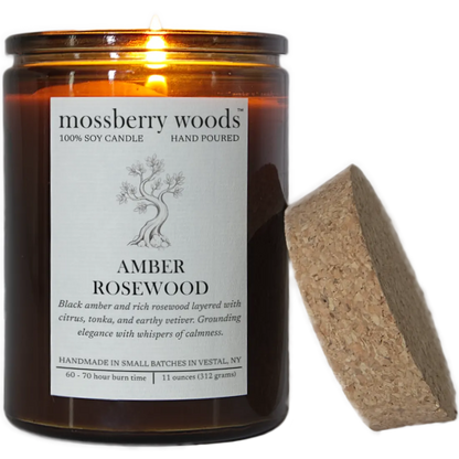 A lit Amber Rosewood scented candle with it's flame illuminating a rustic amber glass container and its cork lid resting on the side. Transparent background.