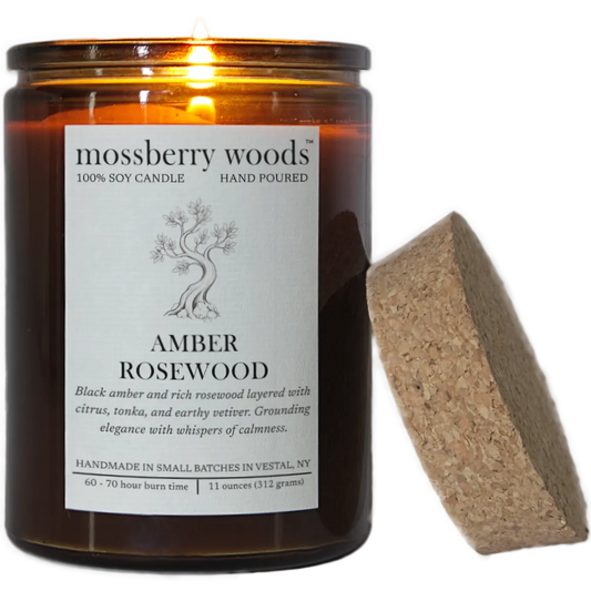 A lit Amber Rosewood scented candle with it's flame illuminating a rustic amber glass container and its cork lid resting on the side. Transparent background.