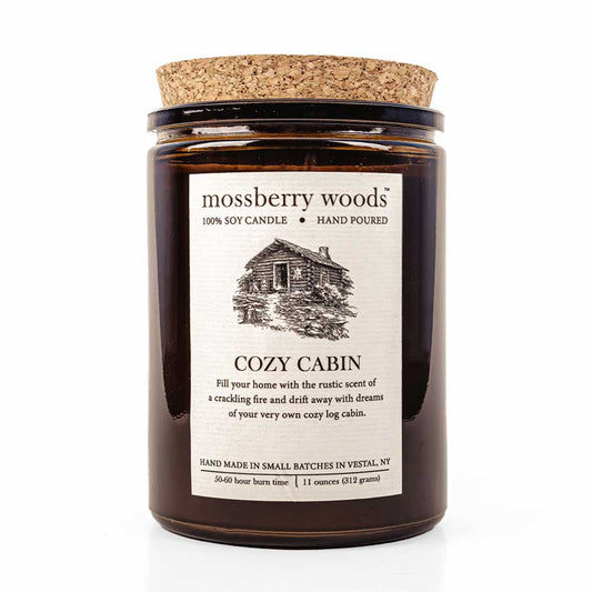 Cozy Cabin  rustic candle in an amber jar with a cork lid showing a log cabin on the label on a white background