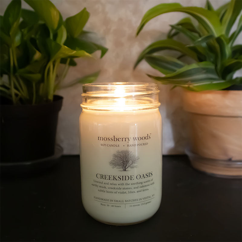Creekside Oasis farmhouse candle lit on wood table with plants in the background