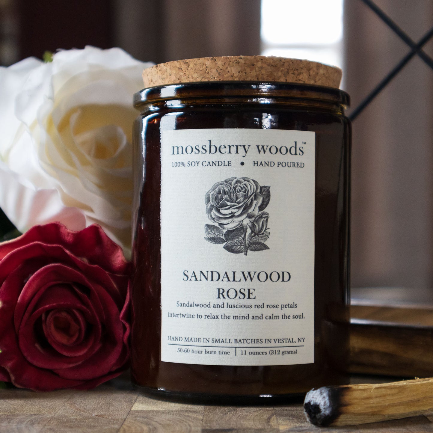 Sandalwood Rose Rustic Candle in an amber jar with a cork lid and a sandalwood stick and roses sitting on a wood table